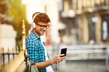 young student with glasses standing on the street against a metal fence and listening music from his...