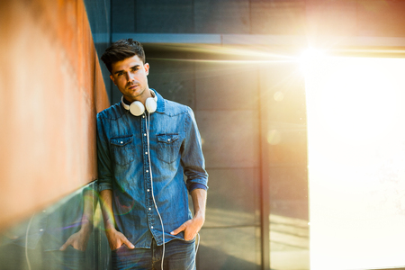 young guy all in jeans outfit, with headsets around his neck, leaning against a wallの写真素材