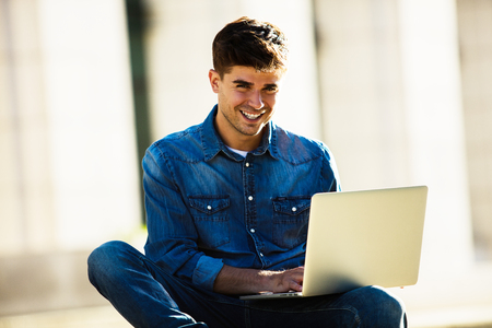 smiling man with laptop in front looking to camera, in front of a big buildingの写真素材