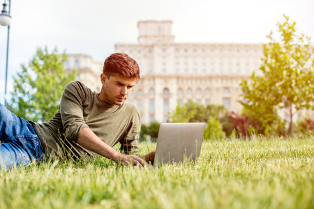 handsome young man, student, lying in grass, looking or working on laptop, outside in park in center of a cityの写真素材