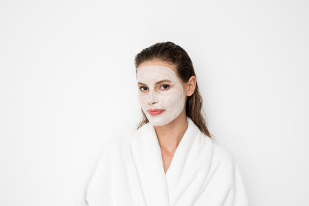 Beautiful young girl with moisturizing facial mask on white background. Photo of girl in bathrobe...