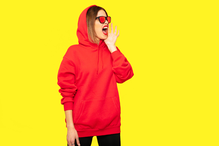 cool hipster girl with sunglasses screeming at someone, or protesting about something, in red hoodieの写真素材