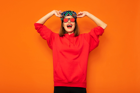 young and cool hipster girl having fun, with colored hat and sunglasses posing with good vibes to camera on orange backgroundの写真素材