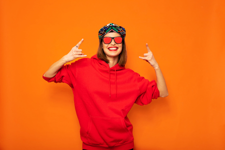 young and cool hipster girl having fun, with colored hat and sunglasses posing with good vibes to camera on orange backgroundの写真素材