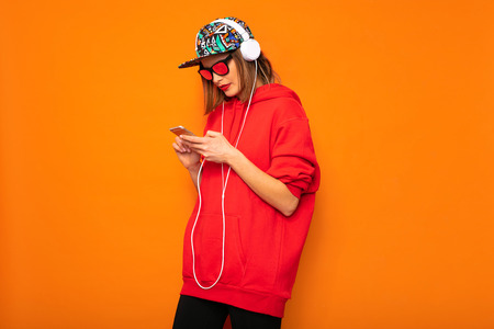 cool hipster girl looking at her cellphone and listening music on headphones, wearing colored hat on orange backgroundの写真素材