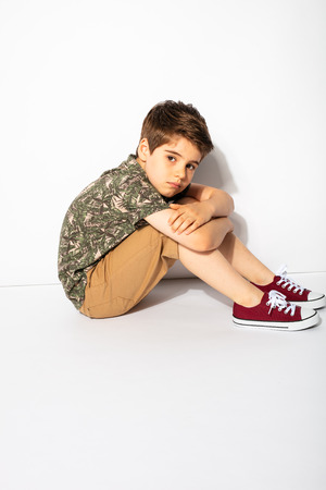 alone little boy sitting down and looking sad to the camera on white backgroundの写真素材