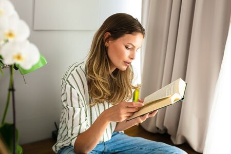 woman reading next to a bright window, relaxing and chilling during the outbreak period. Home...