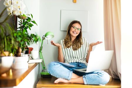 smiling woman working from home with laptop and pappers around her next to a bright window and the houseplantの写真素材