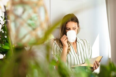 woman reading next to a bright window, relaxing , drinking coffee and chilling during the outbreak period. Home activity for healthy mindの写真素材
