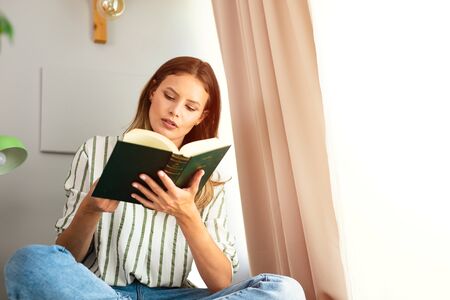 woman reading next to a bright window, relaxing and chilling during the outbreak period. Home activity for healthy mindの写真素材