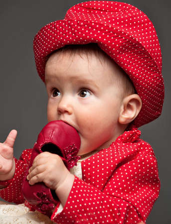 Beautiful baby girl dressed in a red dress eating her own delicious red shoe while looking very surprisedの写真素材