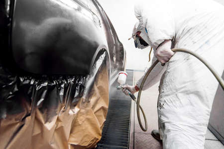 auto worker spraying black paint on a car in an auto garage with protective gearの写真素材