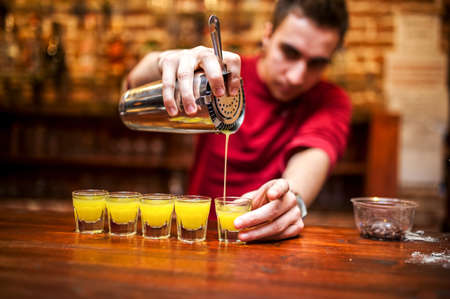 barman mixing and pouring a summer alcoholic cocktail into small glasses on counterの写真素材
