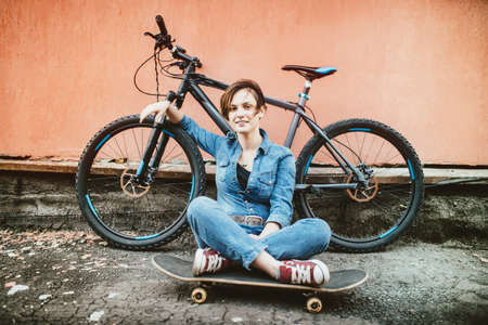 Young sexy brunette girl with short hair standing near vintage bicycle and holding a skateboard, having fun and a good mood while looking in camera and smilingの写真素材