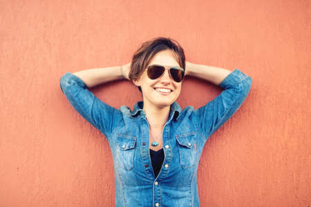 Trendy brunette girl, making face expressions, smiling and laughing against orange background, isolated. Modern lifestyle with beautiful hipster girl with sunglassesの写真素材