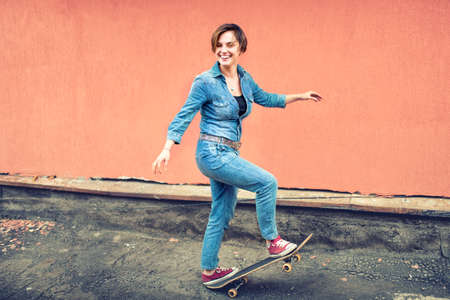 Artsy portrait of a brunette cute girl on a skateboard, laughing and having a good time. Healthy concept of modern life, hipster girl with skateboard on rooftopの写真素材