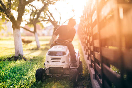 Male riding lawnmower and cutting grass during sunset golden hour. Details of gardening with sunrays