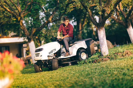lawncare concept - handsome young man trimming grass in garden using tractorの写真素材