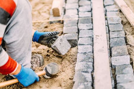 Close up details of construction works with industrial worker placing granite cobblestone blocks on path or alleyの写真素材