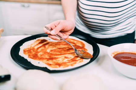 Young caucasian beautiful pregnant woman preparing dough for pizza, pouring tomatoes sauce の写真素材