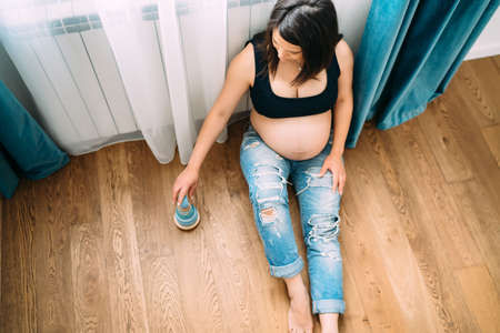 Beautiful pregnant woman enjoying life and playing with future baby toys, sitting on hardwood floorの写真素材