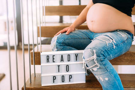 Ready to pop concept - pregnant woman portrait with close up of belly on stairs in homeの写真素材