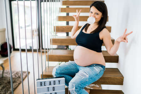 Cheerful portrait of young pregnant woman  sitting on stairs enjoying life. modern lifestyle details - new home and baby comingの写真素材