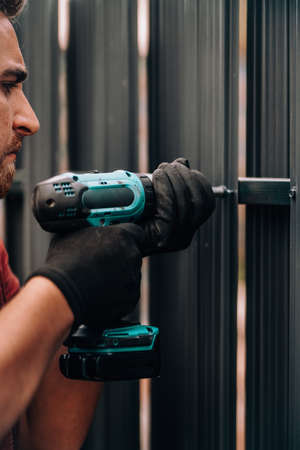 Worker using screwdriver drilling and screwing metal screws into metal fenceの写真素材