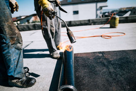 Construction workers insulating rooftop with bituminous membrane. Waterproofing details at construction site. Bituminous membrane waterproofing system details and installation on flat rooftop.の写真素材