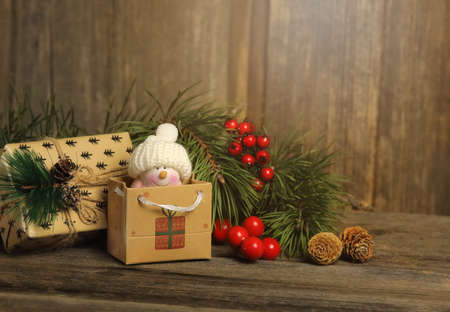 Lovely tiny snowman in the package laying on the wooden background with another present, fir branches, cones and wooden firs are near byの写真素材