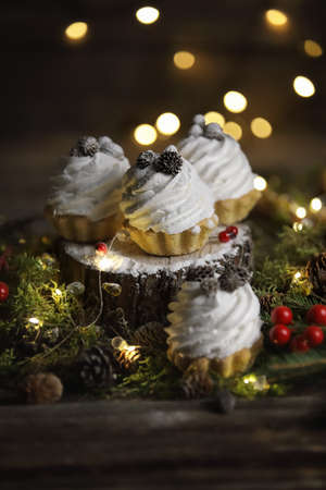 Festive cakes in the form of tartlets with cream on a dark background with fir branches, garland is around tartlets, christmas mood, festive tableの写真素材