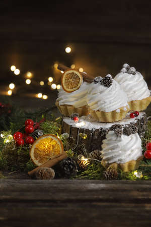 Festive cakes in the form of tartlets with cream on a dark wooden background with moss, cinnamon, orange cones. Garland is around tartlets, christmas mood, festive tableの写真素材