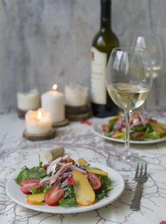 Salad with persimmon, smoked chicken, cherry tomatoes, Camembert cheese and blue cheese sauce. Festive dish on a white wooden table with a glass of white wine. Christmas dinner. Selective focus.の写真素材
