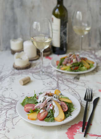 Salad with persimmon, smoked chicken, cherry tomatoes, Camembert cheese and blue cheese sauce. Festive dish on a white wooden table with a glass of white wine. Christmas dinner. Selective focusの写真素材