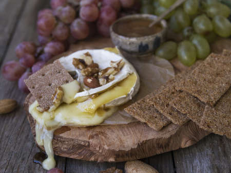 Baked Camembert on wooden board, branch of green and red grapes, rosemary, rye bread, nuts and honey. Close up, copy space, selective focusの写真素材