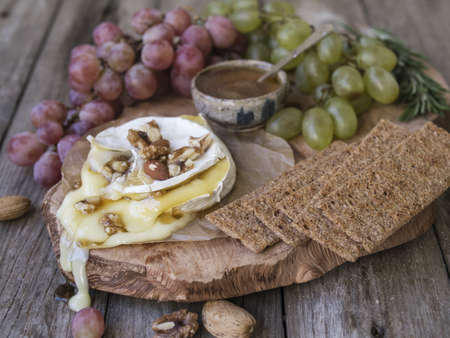 Baked Camembert on wooden board, branch of green and red grapes, rosemary, rye bread, nuts and honey. Close up, copy space, selective focusの写真素材