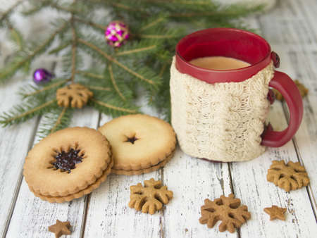Winter morning, cup of coffee with knitted cup holders, gingerbread cookies with fir tree branches decorated with spruce balls on white wooden background.Breakfast.Ideal for winter couple in loveの写真素材