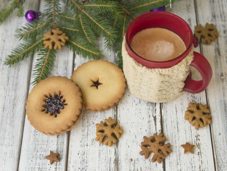 Winter morning, cup of coffee with knitted cup holders, gingerbread cookies with fir tree branches decorated with spruce balls on white wooden background.Breakfast.Ideal for winter couple in loveの写真素材