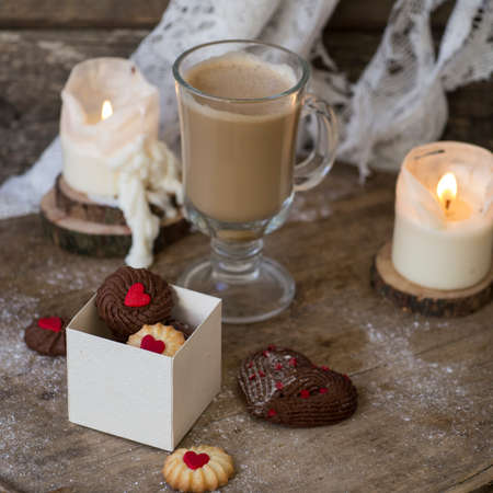 Heart shaped Viennese chocolate and vanilla Valentine's Day cookies with a glass cup of coffee with milk on a wooden background with candles. Valentine's day background. Selective focus, close upの写真素材