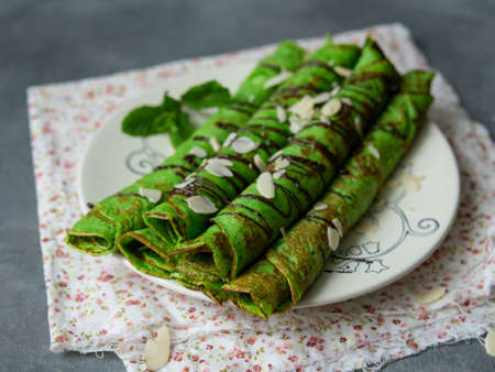 Mint thin crepes of green color, twisted into tubes, with chocolate sauce and almond petals on a white plate on gray background with floral fabric. Exquisite breakfast. Close up, selective focus..の写真素材