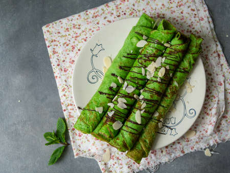 Mint thin crepes of green color, twisted into tubes, with chocolate sauce and almond petals on a white plate on gray background with floral fabric. Exquisite breakfast. Close up, selective focus.の写真素材