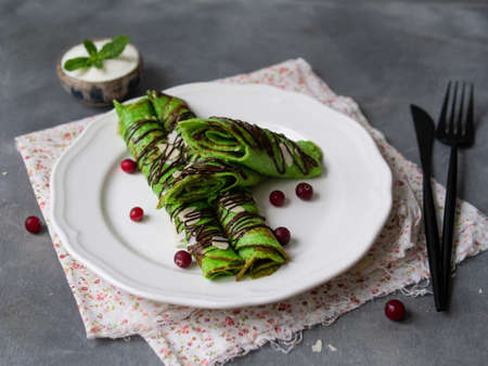 Mint thin crepes of green color, twisted into tubes, with chocolate sauce, sour Cream, mint leaf and almond petals on a white plate on gray background with floral fabric. Breakfast. Selective focus.の写真素材