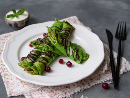 Mint thin crepes of green color, twisted into tubes, with chocolate sauce, sour Cream, mint leaf and almond petals on a white plate on gray background with floral fabric. Breakfast. Selective focus.の写真素材