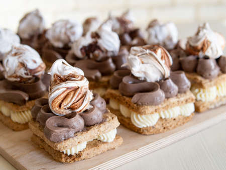 Portion cakes of meringue and cocoa butter cream in square form on a wooden stand, a piece of cake on a light wooden background. Restaurant serving. Selective focus, close upの写真素材