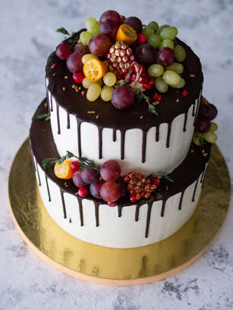 Two-tiered white cake with fresh fruits - pomegranate, green and red grapes, kumquat, cranberries, thyme sprigs - and chocolate drips. Autumn cake. birthday cake, anniversary. Close upの写真素材