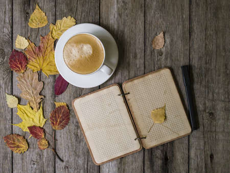 Autumn background on a wooden table with autumn dry colored leaves, white cup of coffee and a notepad with a pencil. Cozy warm mockup. Autumn mood. Top view, close up, copy space, flat lay.の写真素材
