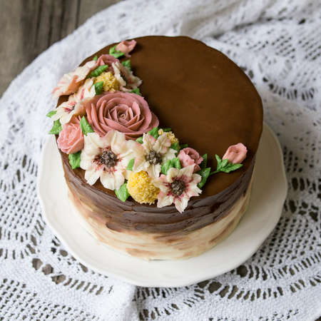 Brown cream cake decorated with buttercream flowers - roses, lilies on wooden background with lace fabric. Close up, top viewの写真素材