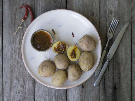 Boiled potatoes in their skins. whole with red pepper, salt and spicy sauce on white plate, old wooden table, rustic style. Close up, copy space. Top view.の写真素材