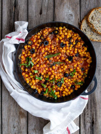 Baked chickpea with vegetables, olives and sun-dried tomatoes in iron pan, frying pan on wooden table. Vegan vegetarian lunch, paleo diet, healthy dinner. Top view, copy space.の写真素材