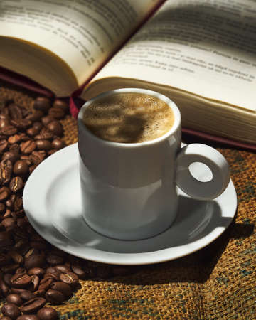 Cup of coffee with foam. Coffee beans, an open book, hemp bag background and lighting with shadows.の写真素材